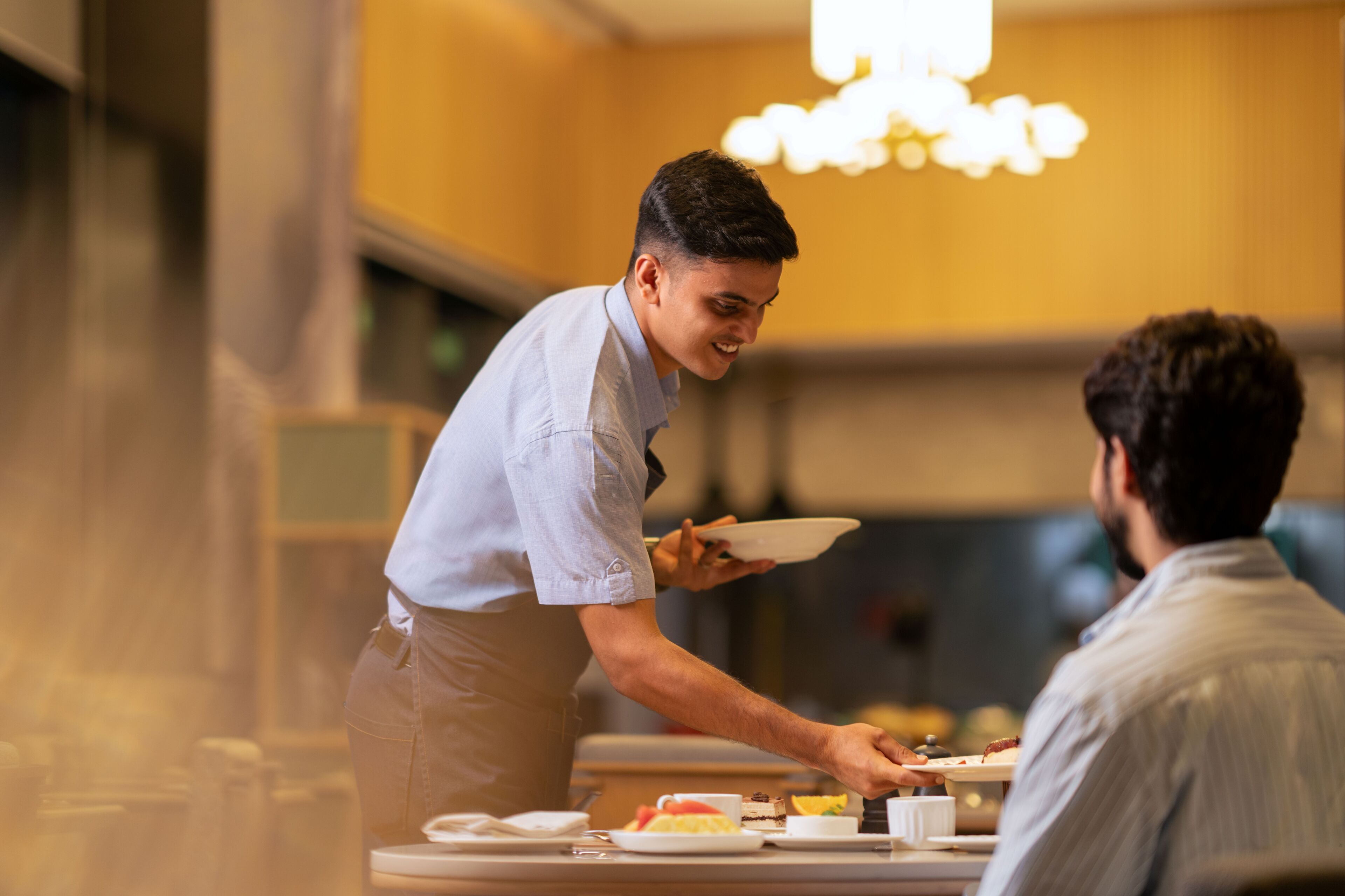 A waiter serving food to a guest in a well-lit dining area with a chandelier.