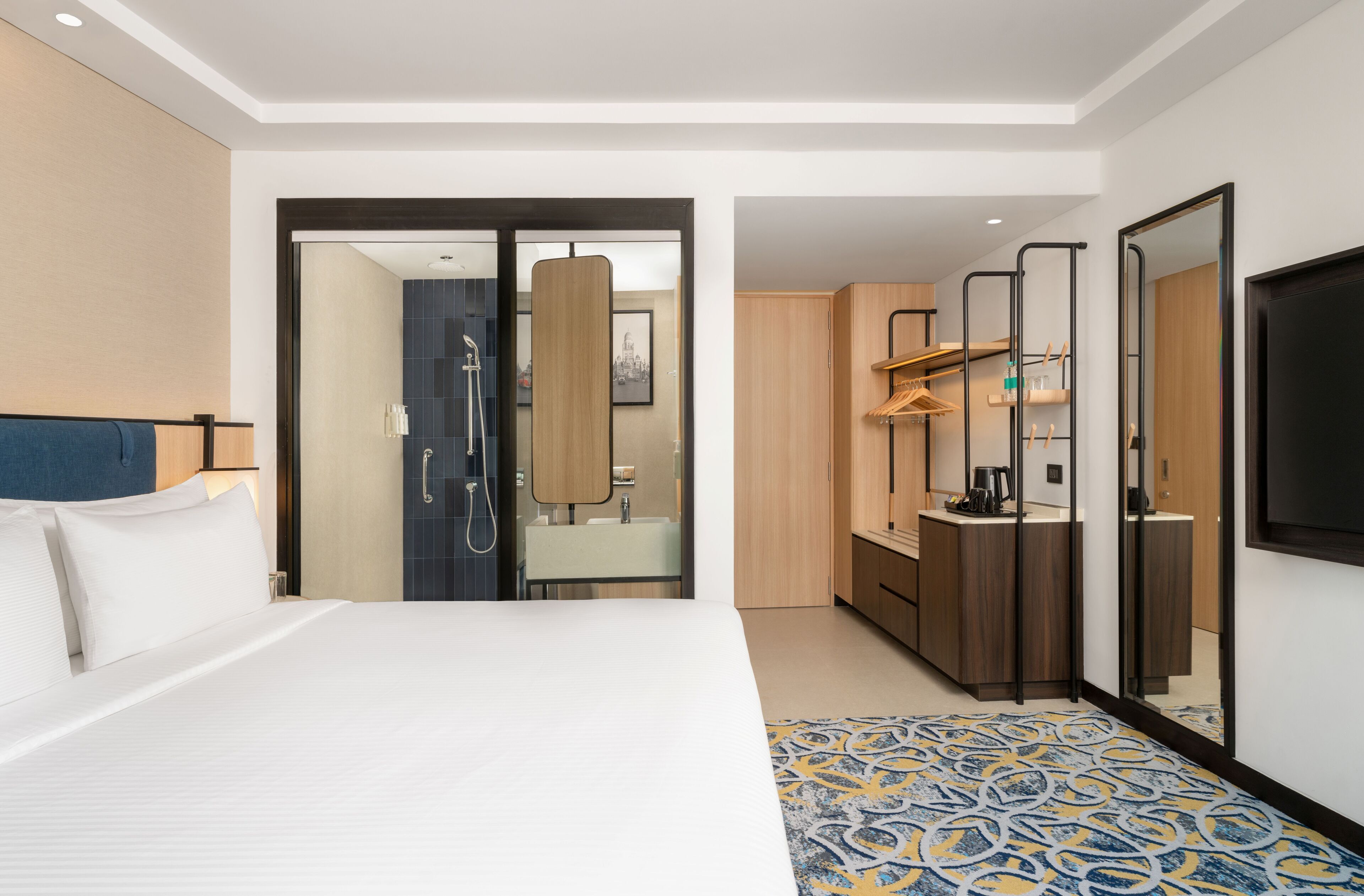 A hotel room with a large bed, a bathroom with a mirror and sink, a small kitchenette, and a TV mounted on the wall.