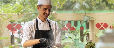 Un cocinero con uniforme blanco y delantal negro cocina al aire libre, con varios cuencos y verduras sobre la encimera.