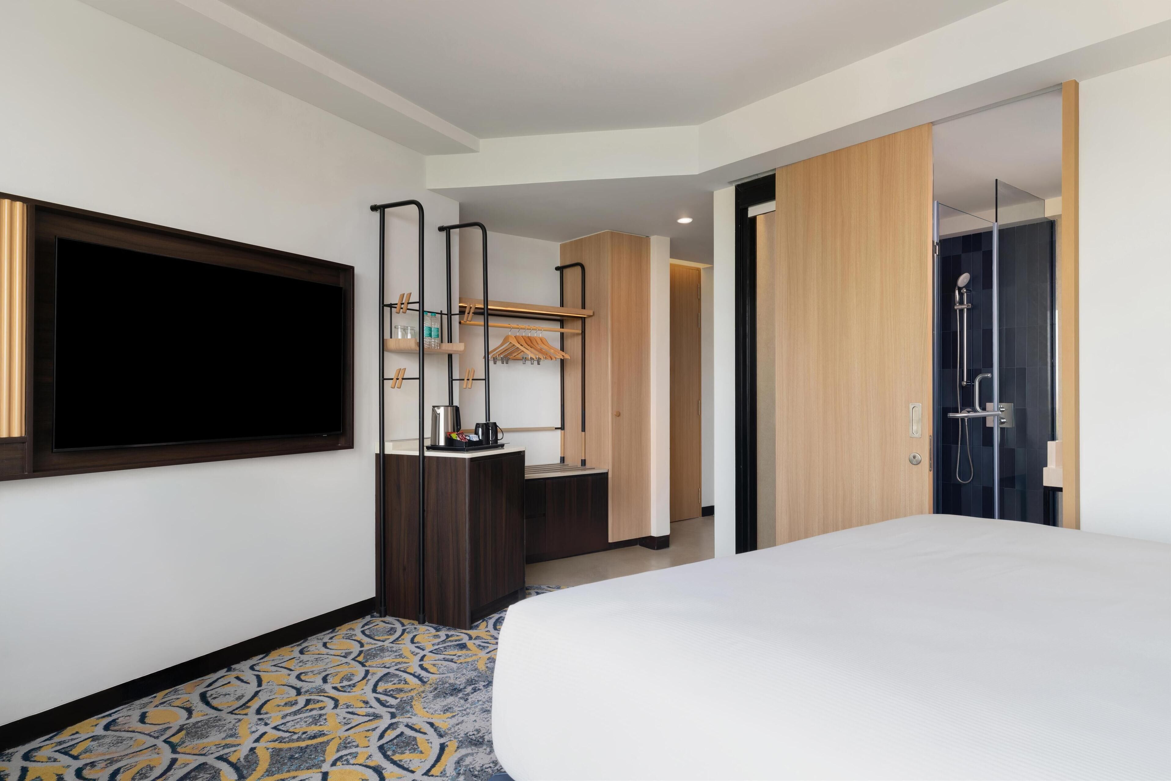 Een hotelkamer met een groot bed, een aan de muur bevestigde televisie, een garderobe, een spiegel, een klein bureau met een lamp en een douche.