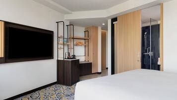 Een hotelkamer met een groot bed, een aan de muur bevestigde televisie, een garderobe, een spiegel, een klein bureau met een lamp en een douche.