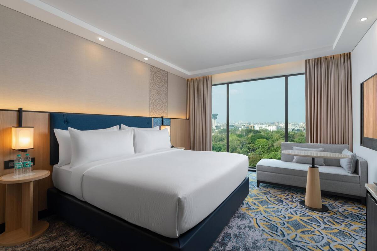 Habitación de hotel con una cama grande, un sofá, una mesita de noche con lámpara y vistas a la ciudad.