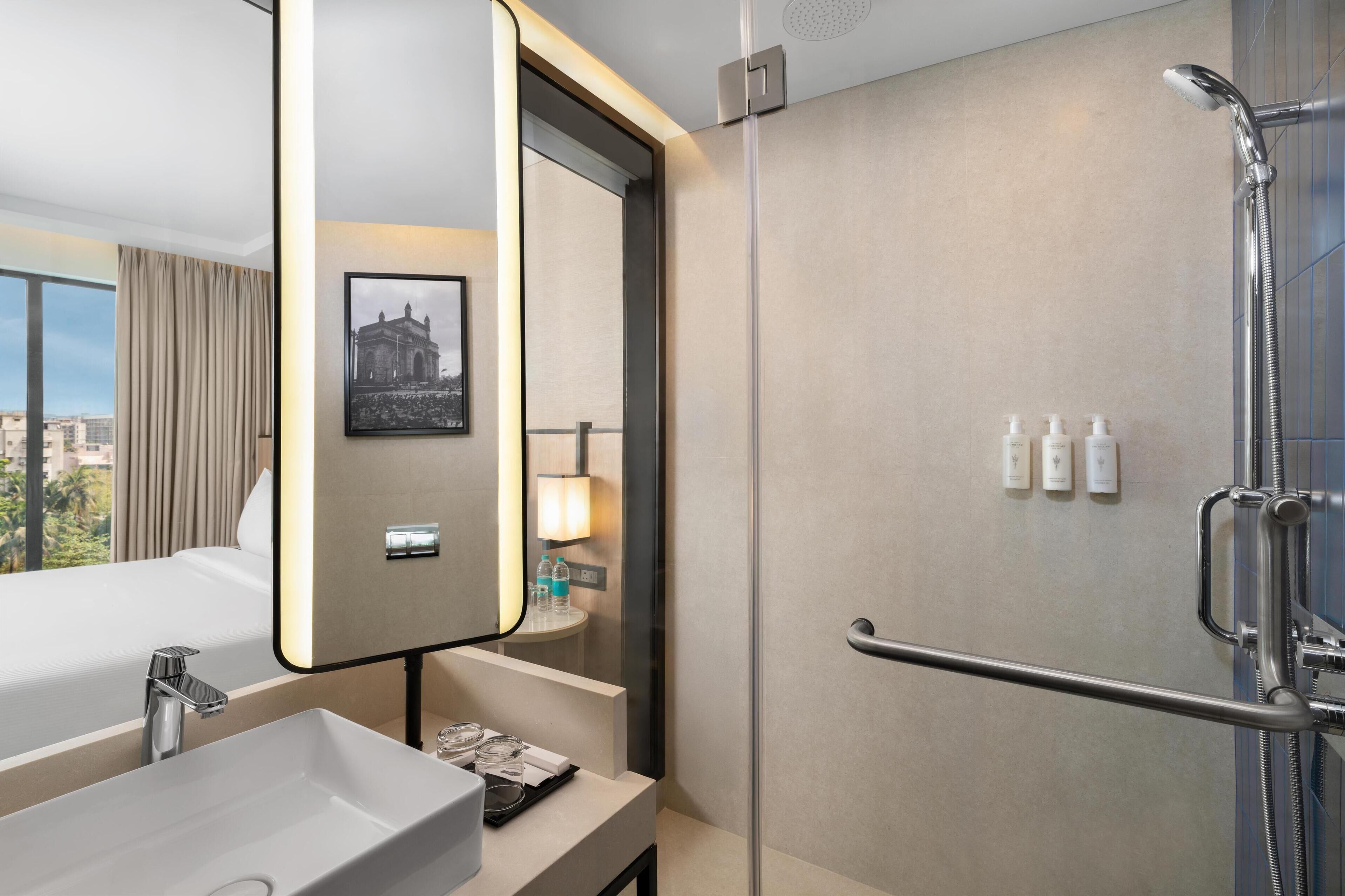A modern hotel bathroom with a large mirror, a sink, a shower, and a view of the city.