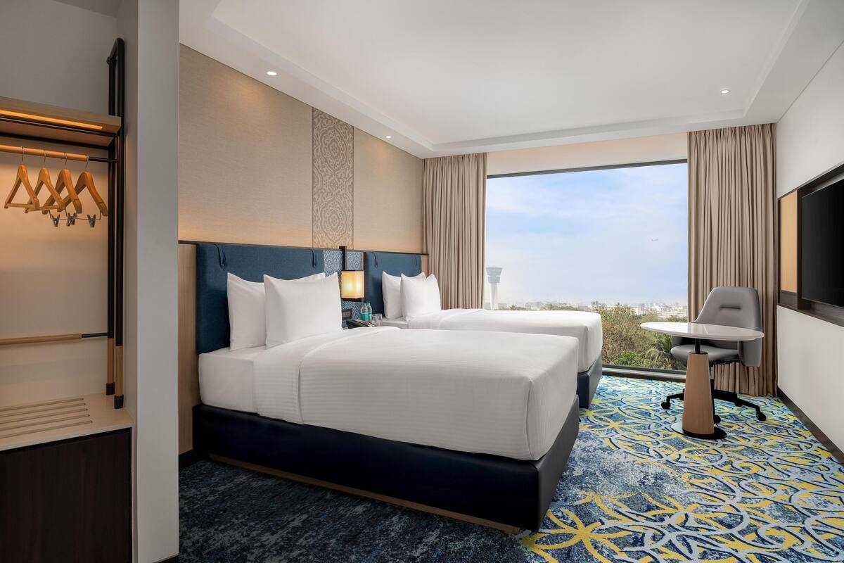 Habitación de hotel con dos camas, un escritorio y un ventanal con vistas a la ciudad.