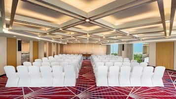 Un salón de conferencias con filas de sillas blancas, una alfombra roja estampada y un techo con diseño geométrico.