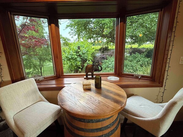 A corner seating area with a round wooden table, two chairs, and a view of greenery outside.
