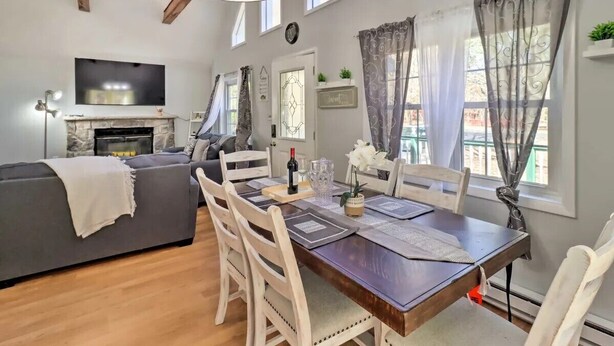 A dining area with a wooden table set for four, surrounded by white chairs. A fireplace is visible in the background, and there is a television mounted on the wall.