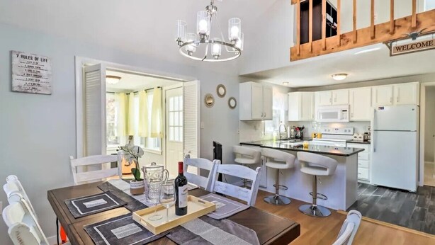 Un coin repas avec une table en bois, des chaises blanches, un lustre et une cuisine en arrière-plan.