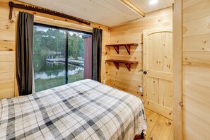 A wooden cabin room with a bed, a window with curtains, a door, and a small shelf.