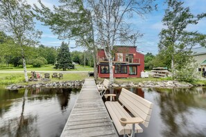 Een houten steiger die leidt naar een bankje aan een rustig meer, omringd door bomen en een rood gebouw met terras.
