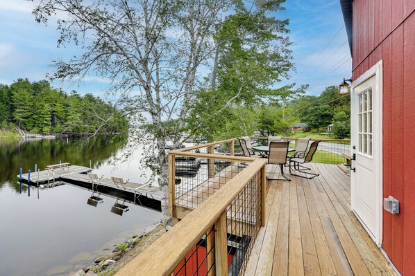 Une terrasse en bois offrant une vue sur un lac, un bâtiment rouge et des meubles d’extérieur.