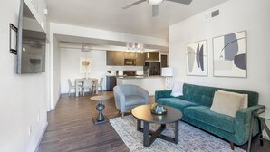 A modern living room with a green sofa, a round coffee table, and a grey armchair. There is a dining area with a table and chairs, and a kitchen in the background. - Landing | Stylish 2BD, Gym (Glendale)