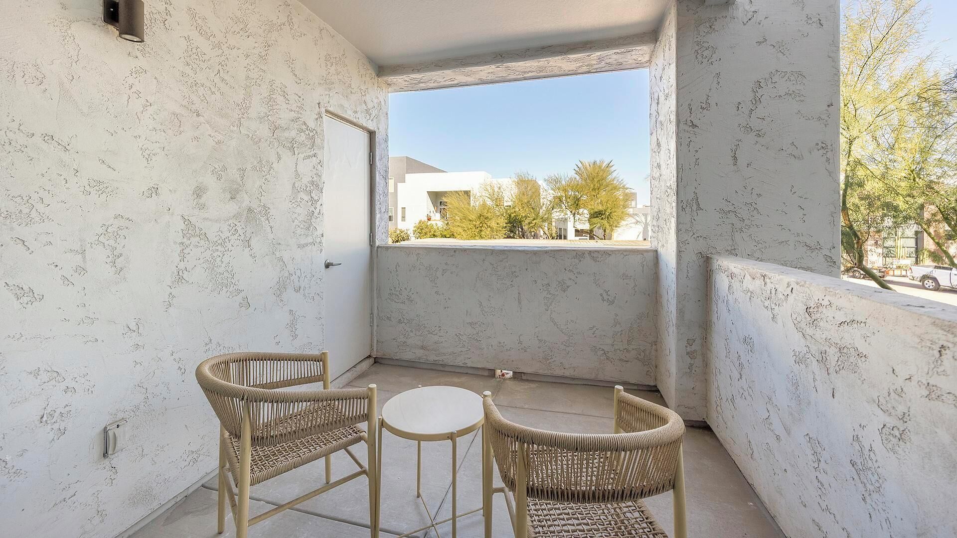A balcony with two wicker chairs and a small round table, white textured walls, and a view of buildings and trees.