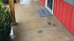 A covered porch with a red and gray building, a metal doormat, and a wooden pillar.