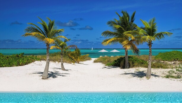 A beach with a clear blue pool, palm trees, and a sailboat in the distance.