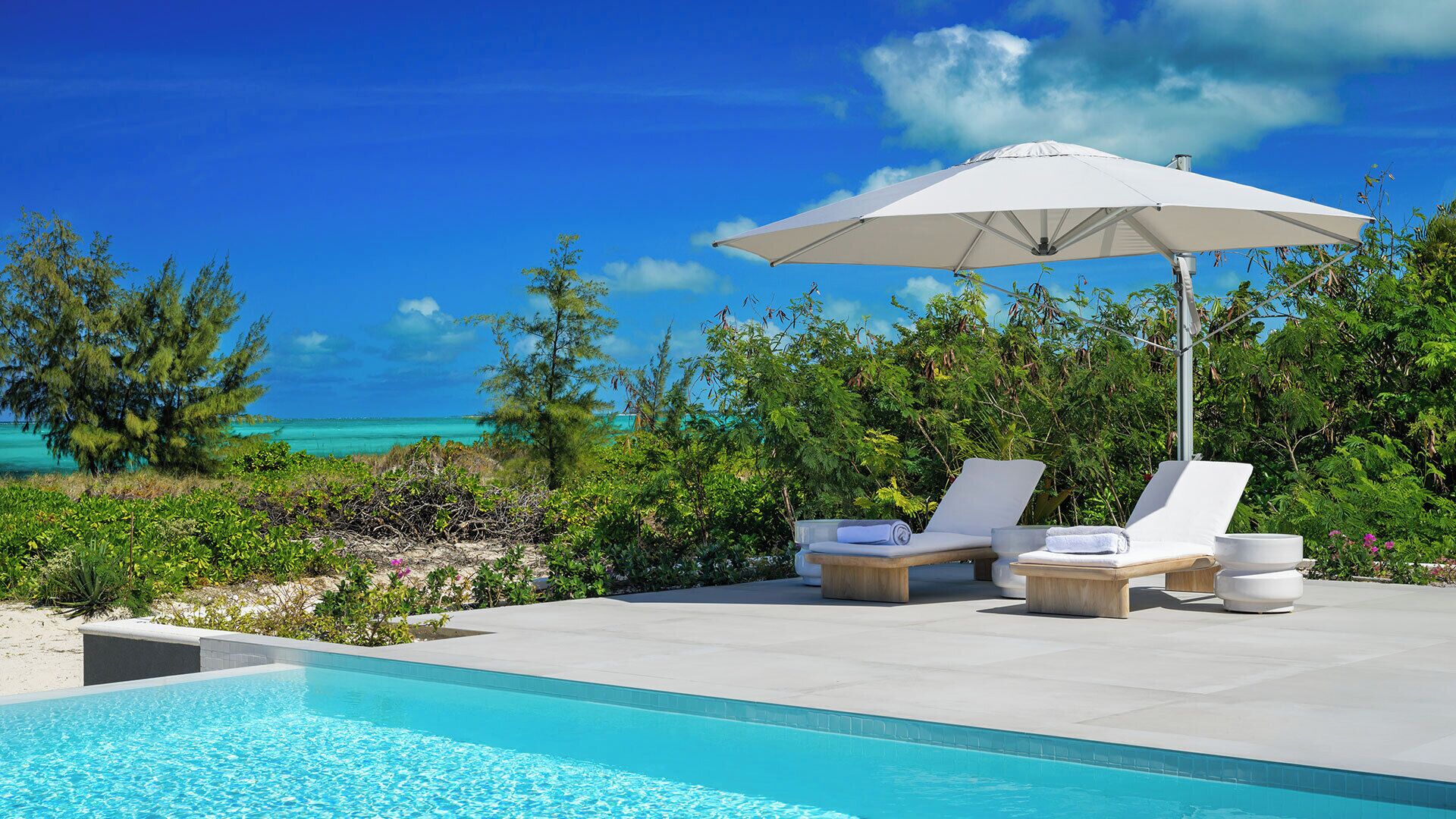 A poolside area with lounge chairs and an umbrella, overlooking a clear blue sea and lush greenery.