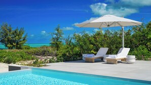 A poolside area with lounge chairs and an umbrella, overlooking a clear blue sea and lush greenery.