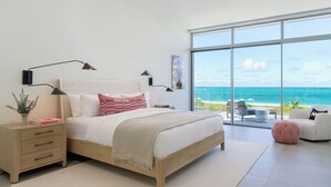 A modern bedroom with a large window overlooking the ocean, a wooden bed, bedside tables, and a small seating area.