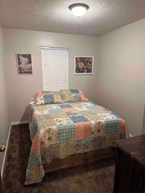 Een slaapkamer met een bed, een raam met jaloezieën en twee foto’s in lijsten aan de muur.