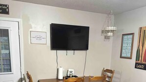 A dining area with a wooden table and chairs, a flat-screen TV mounted on the wall, a chandelier, and a door with a doormat.