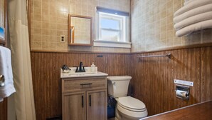 A bathroom with wooden paneling, a sink with a mirror, a toilet, and a towel rack. - Double Queen + Twin | Motel Suite Near Wellsboro (Wellsboro)