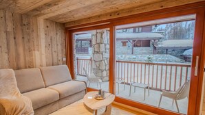 Une pièce chaleureuse avec un plafond en bois, un canapé beige, une table ronde surmontée d’un plateau en marbre, et une vue sur un bâtiment en pierre et un paysage enneigé, visible à travers de grandes portes vitrées.