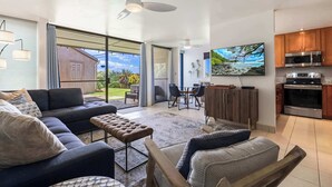 A modern living room with a sofa, ottoman, and a dining area with chairs and a table. There is a kitchen with modern appliances and wooden cabinets.