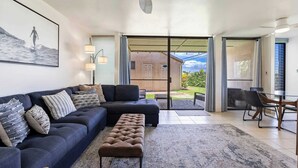 A modern living room with a dark blue sofa, a brown ottoman, and a glass dining table. A large window provides a view of the outdoors.