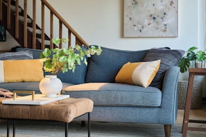 Una sala de estar con un sofá gris, un reposapiés marrón y un jarrón blanco con plantas verdes.