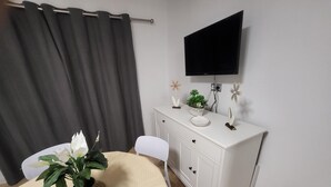 A room with a dining table, a white cabinet, a flat-screen TV mounted on the wall, and a curtain.