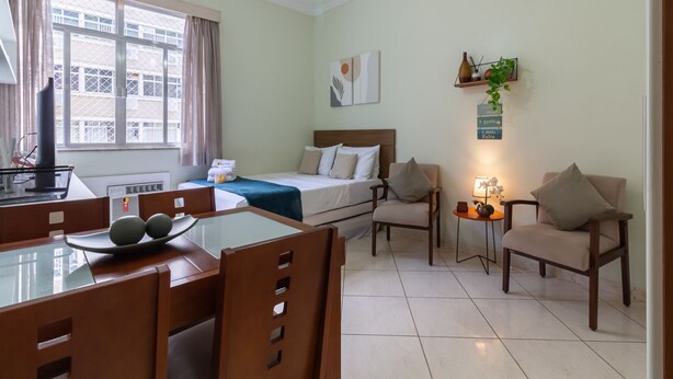 A well-lit room with a bed, a desk with a computer, a dining table with chairs, and a small side table with a lamp. - Studio 5min From Copacabana Beach Mvc901 (Rio de Janeiro)