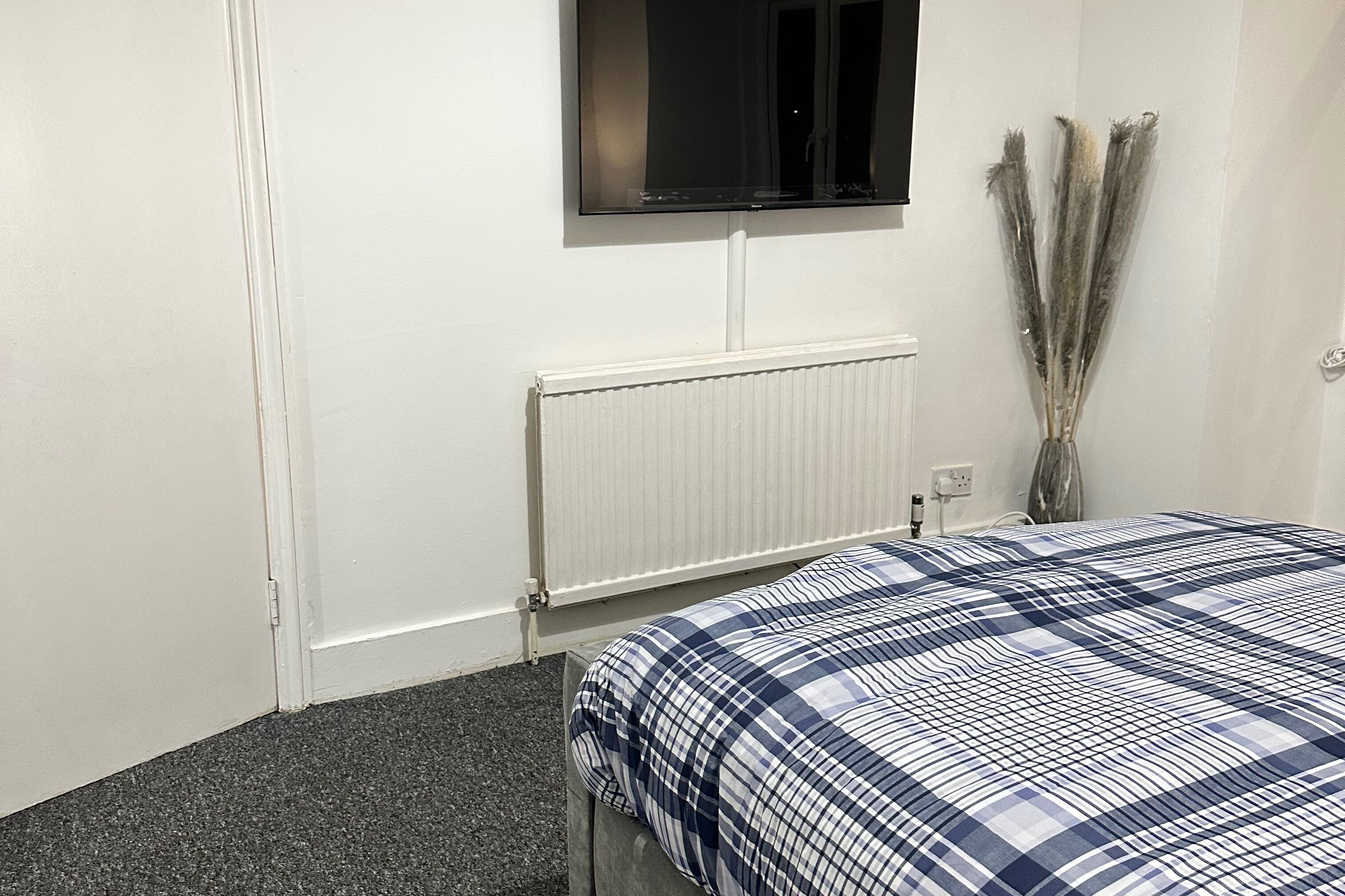 A bedroom with a bed, a radiator, a wall-mounted TV, and a vase with dried plants.