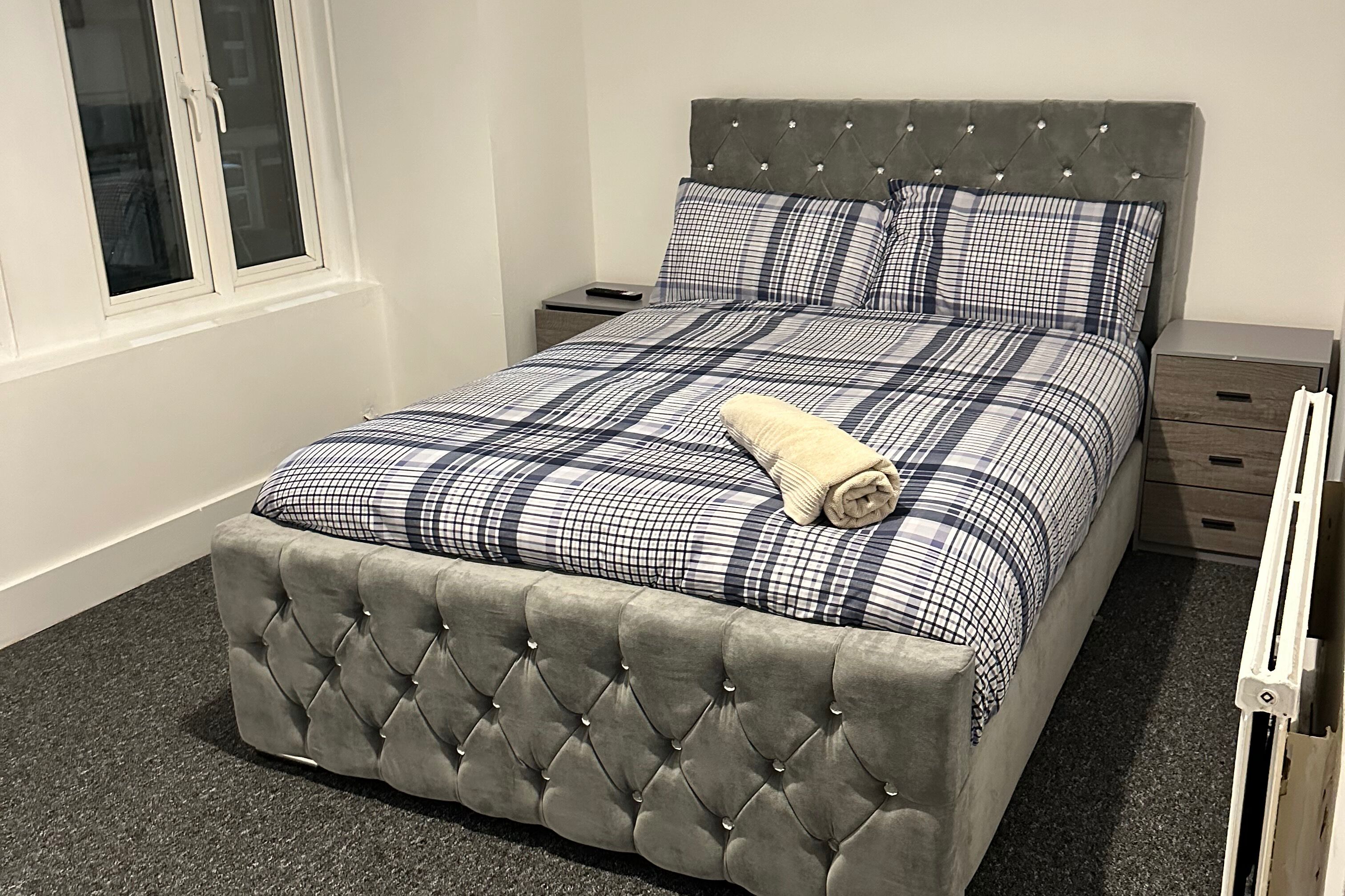 A neatly made bed with a tufted headboard and a plaid bedspread, accompanied by a bedside table and a drawer unit.