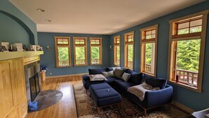 A living room with a blue sofa, a fireplace, and large windows with a view of greenery. - Trout Lake Treehouse - A large, splendid, and private mountain home! (Trout Lake)
