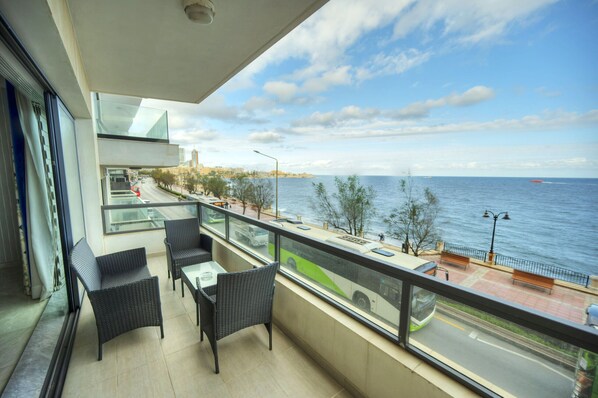 A balcony with wicker furniture, a glass railing, and a view of the sea and cityscape.