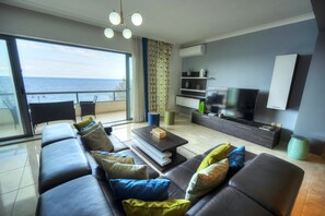 Ein modernes Wohnzimmer mit einer großen Couch, einem Couchtisch und einem Flachbildfernseher. Ein Balkon bietet Meerblick.