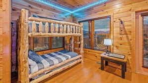 Une chambre dans un chalet en bois, avec un lit superposé, une petite table avec une lampe et une fenêtre avec des stores.
