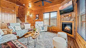Un intérieur de chalet chaleureux avec une cheminée, des murs en bois, un ventilateur de plafond et une grande fenêtre.