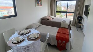 Habitación de hotel con una mesa de comedor preparada para cuatro, dos camas, un ventanal con vistas y un ventilador de techo.