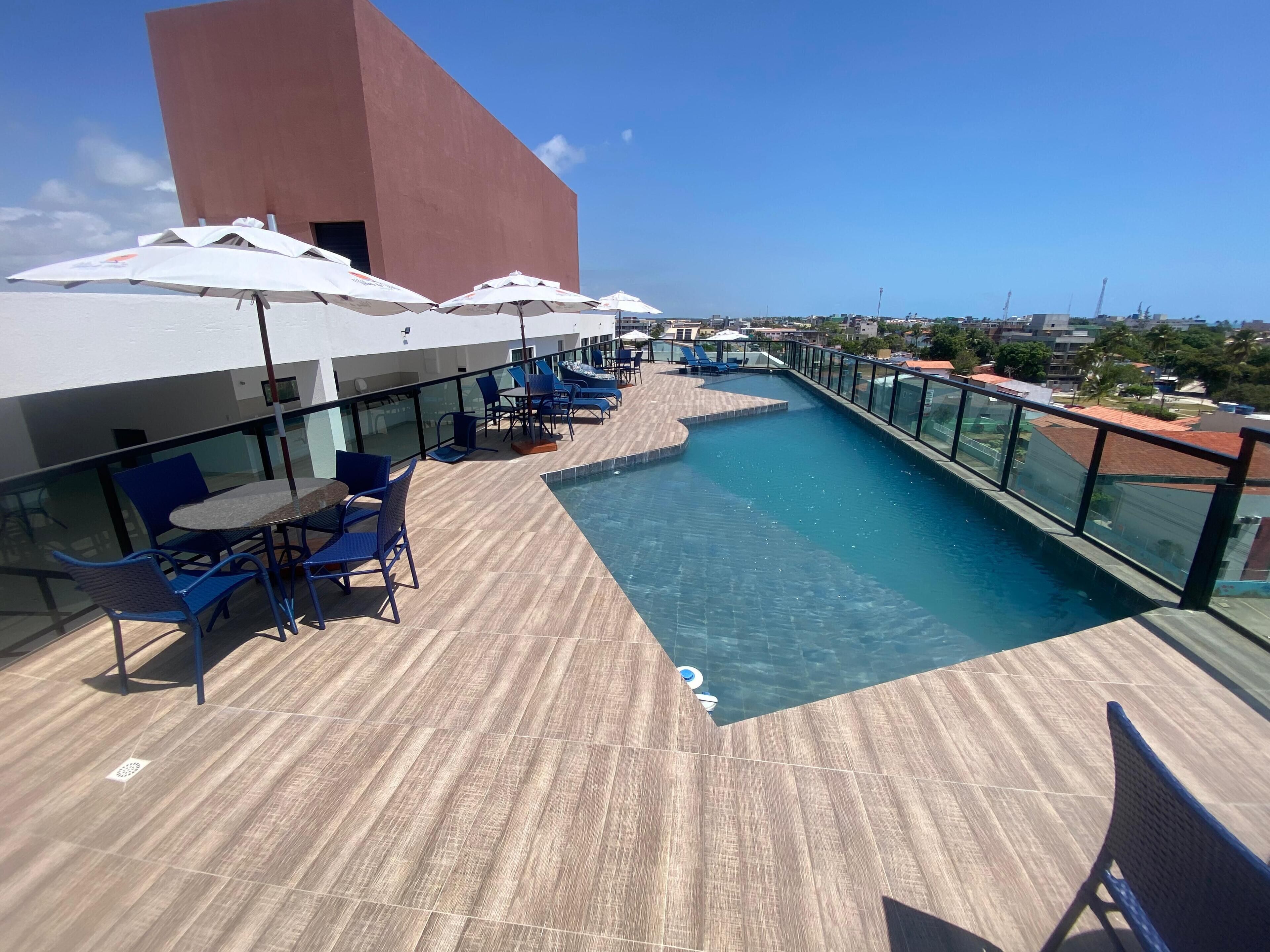 A rooftop pool area with blue lounge chairs and umbrellas, a clear view of the cityscape, and a modern building in the background.