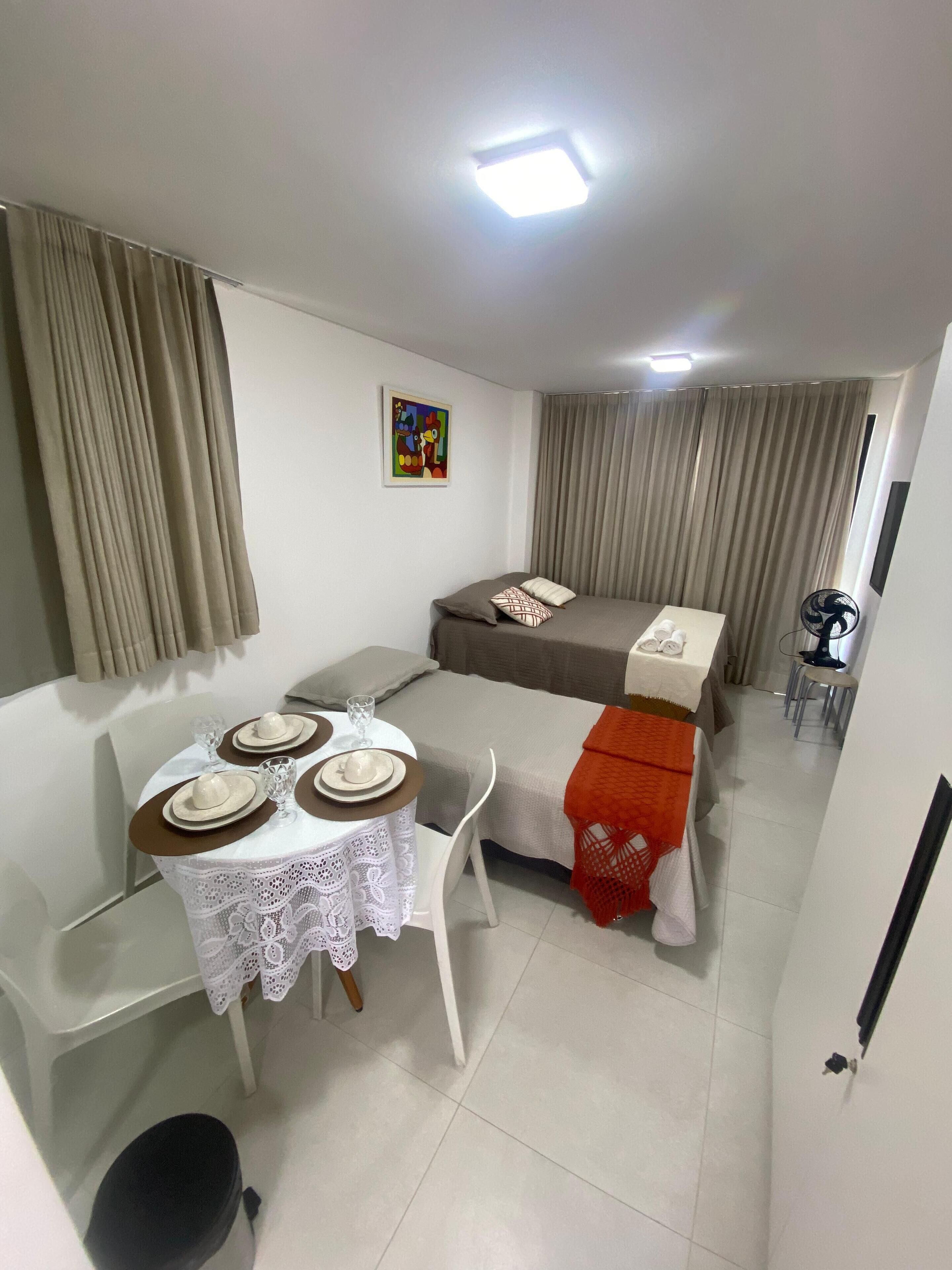 A hotel room with a bed, a dining table set for two, a small table with a decorative tablecloth, and a ceiling light.