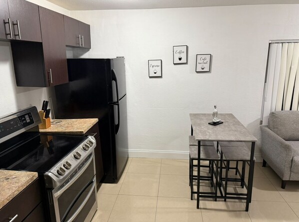 A compact kitchen with dark cabinets, a black refrigerator, a stove, and a small dining table with chairs. There are three framed pictures on the wall. - Miami Central Rest Pad for 2 (Miami)