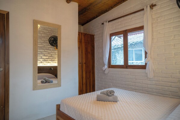 A bedroom with a bed, a mirror, a window with curtains, and a wooden ceiling.