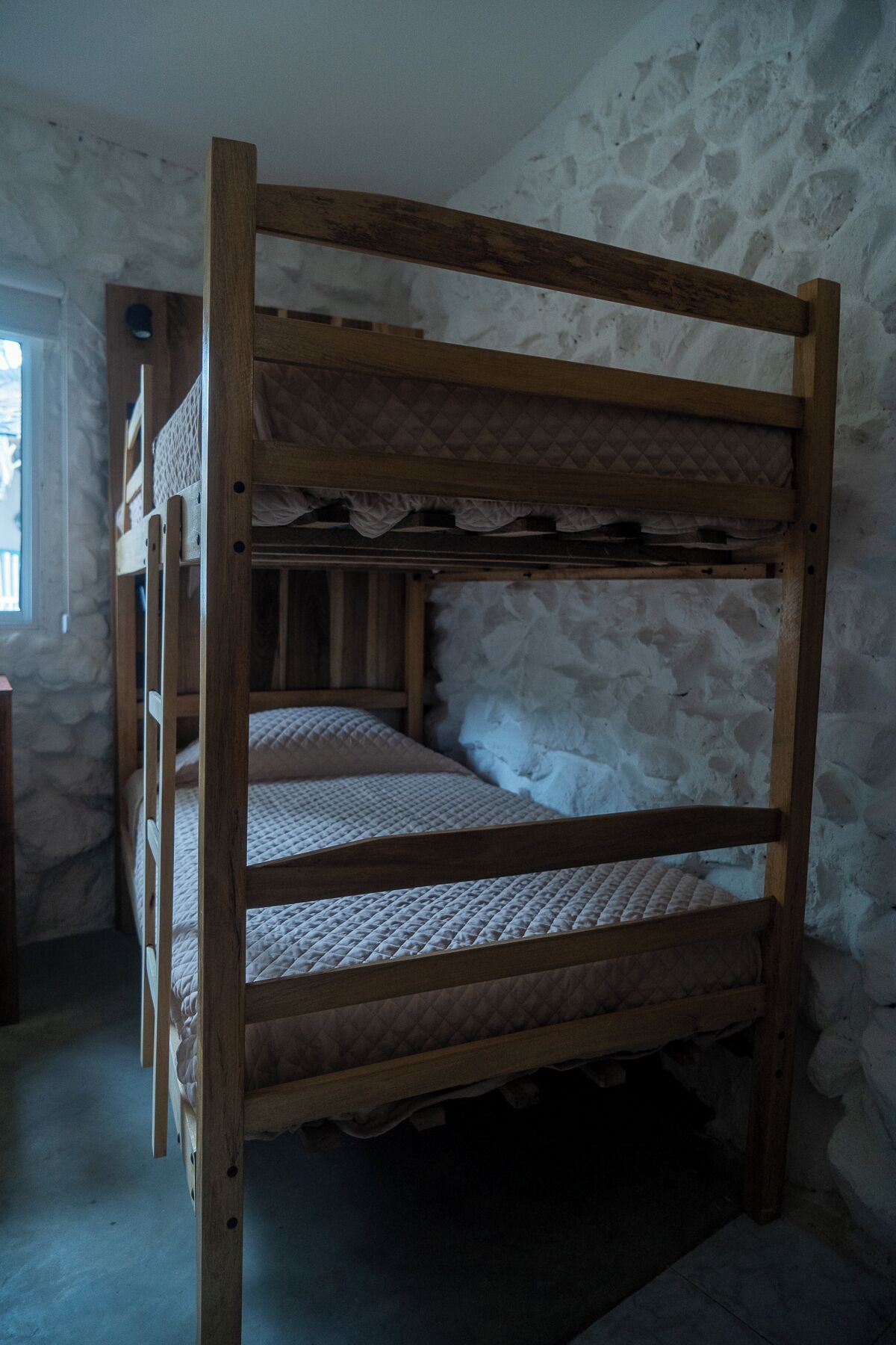 A bunk bed with a wooden frame and quilted bedding in a room with stone walls and a window.