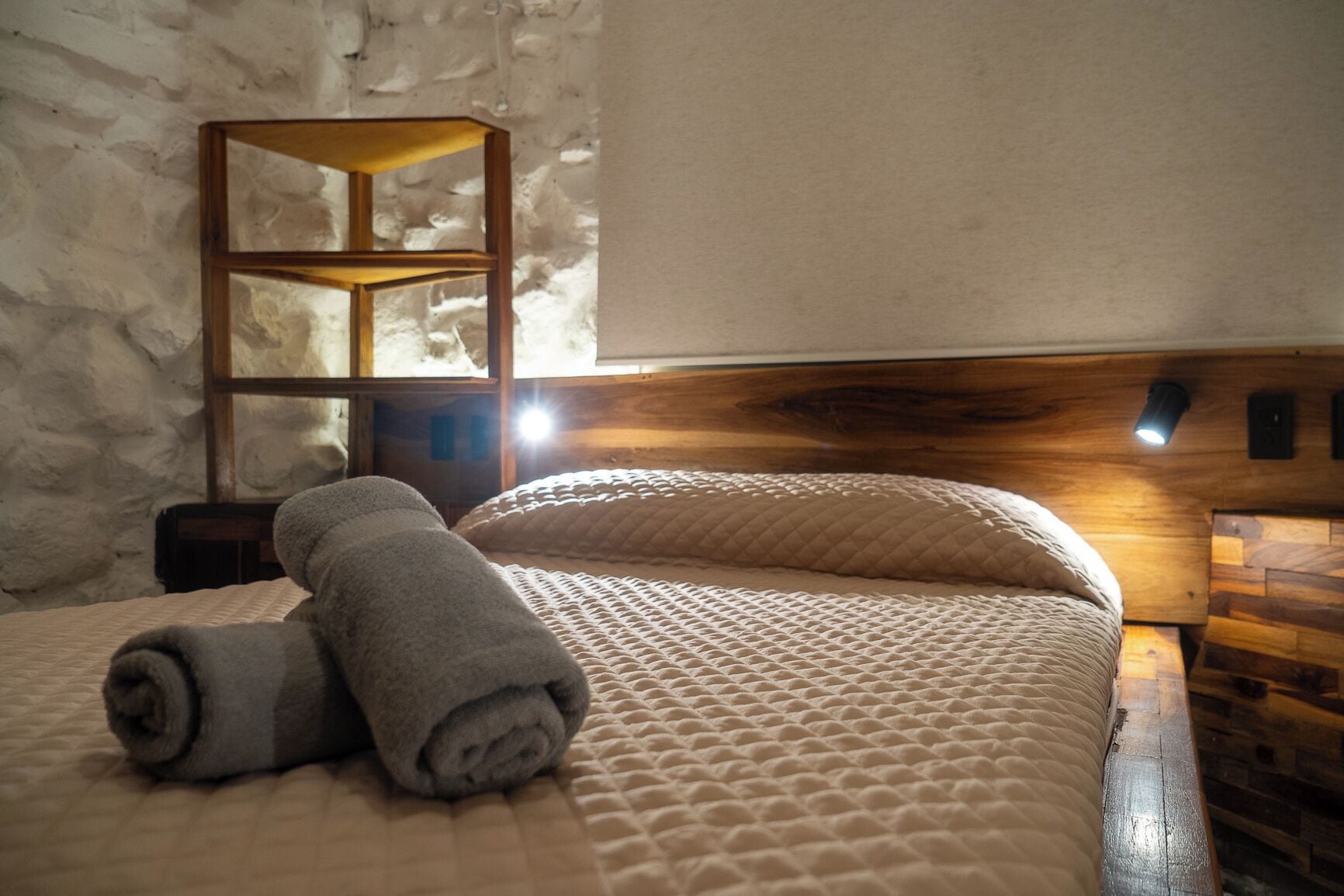 A neatly made bed with a rolled towel, a wooden headboard, and a wall-mounted shelf.