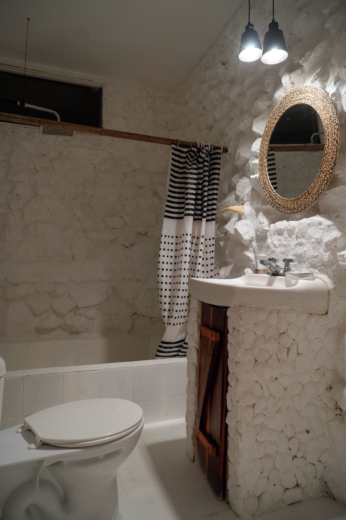 A bathroom with a stone wall, a white sink, a striped shower curtain, and a round mirror.