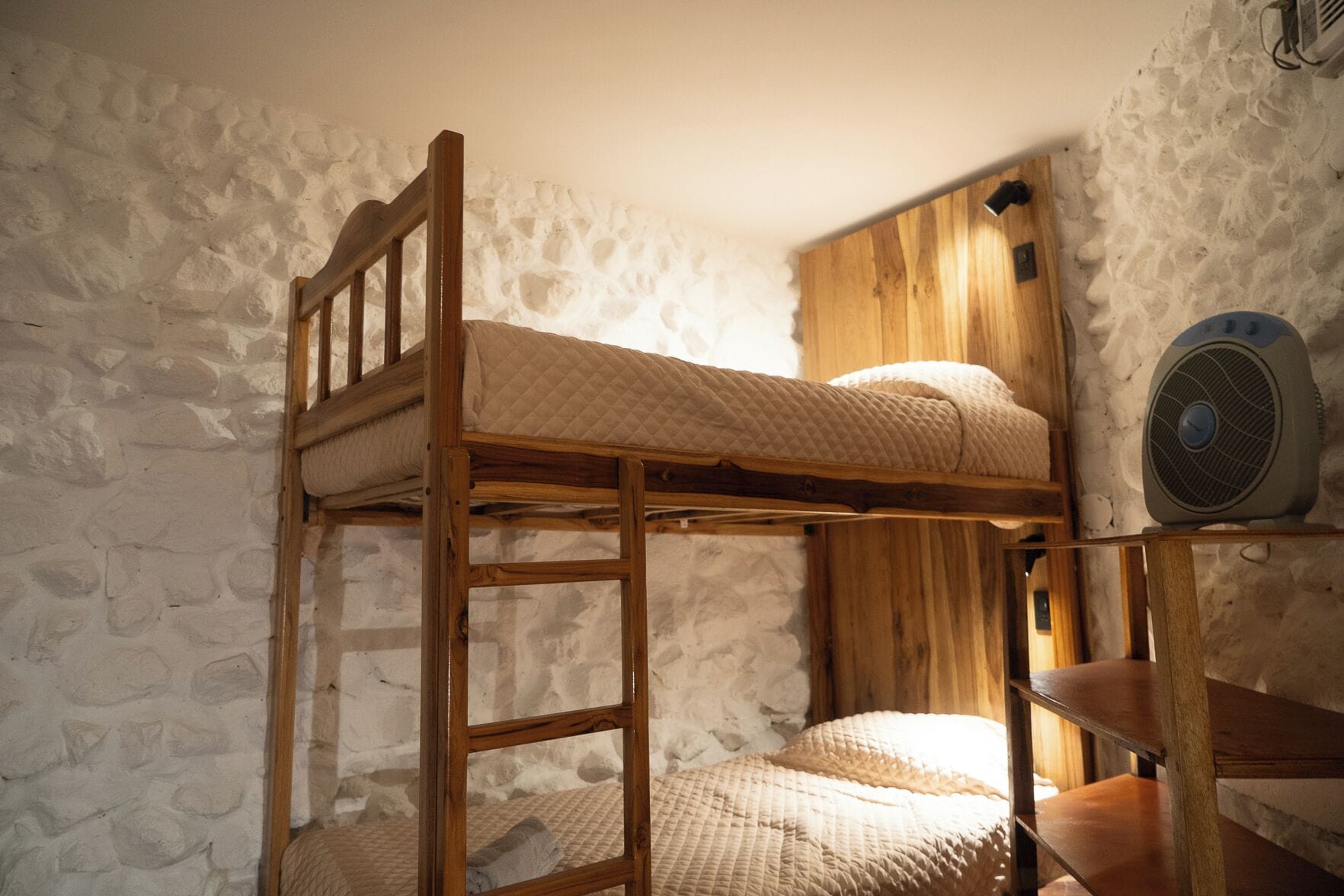 A bunk bed with a wooden ladder, a small table, and a fan against a stone wall.