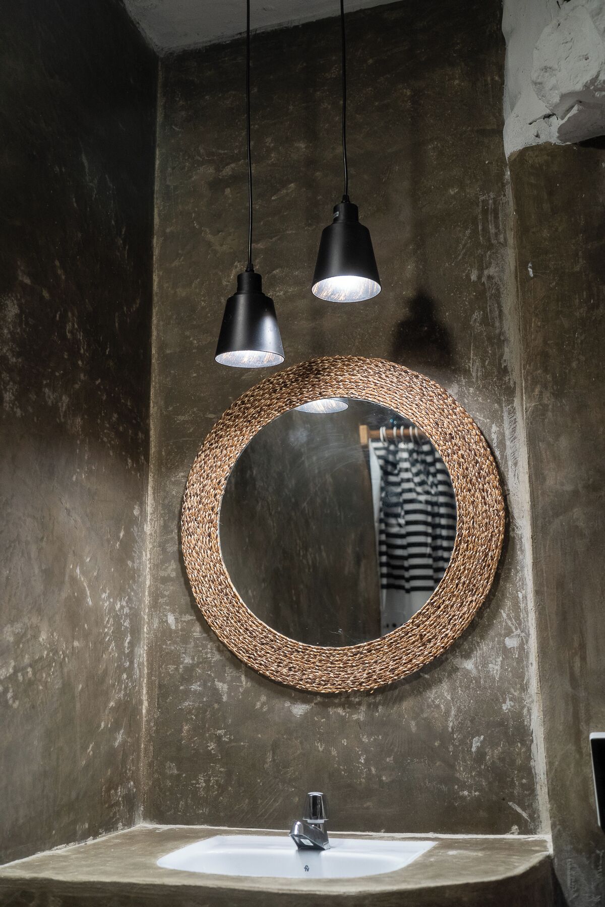 A bathroom with a round mirror surrounded by a woven frame, a sink, and two pendant lights.