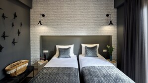 Ein Hotelzimmer mit zwei Betten, einer Wand mit Ziegeloptik, Vogelbilder an der Wand und einem kleinen Beistelltisch mit einer Flasche.