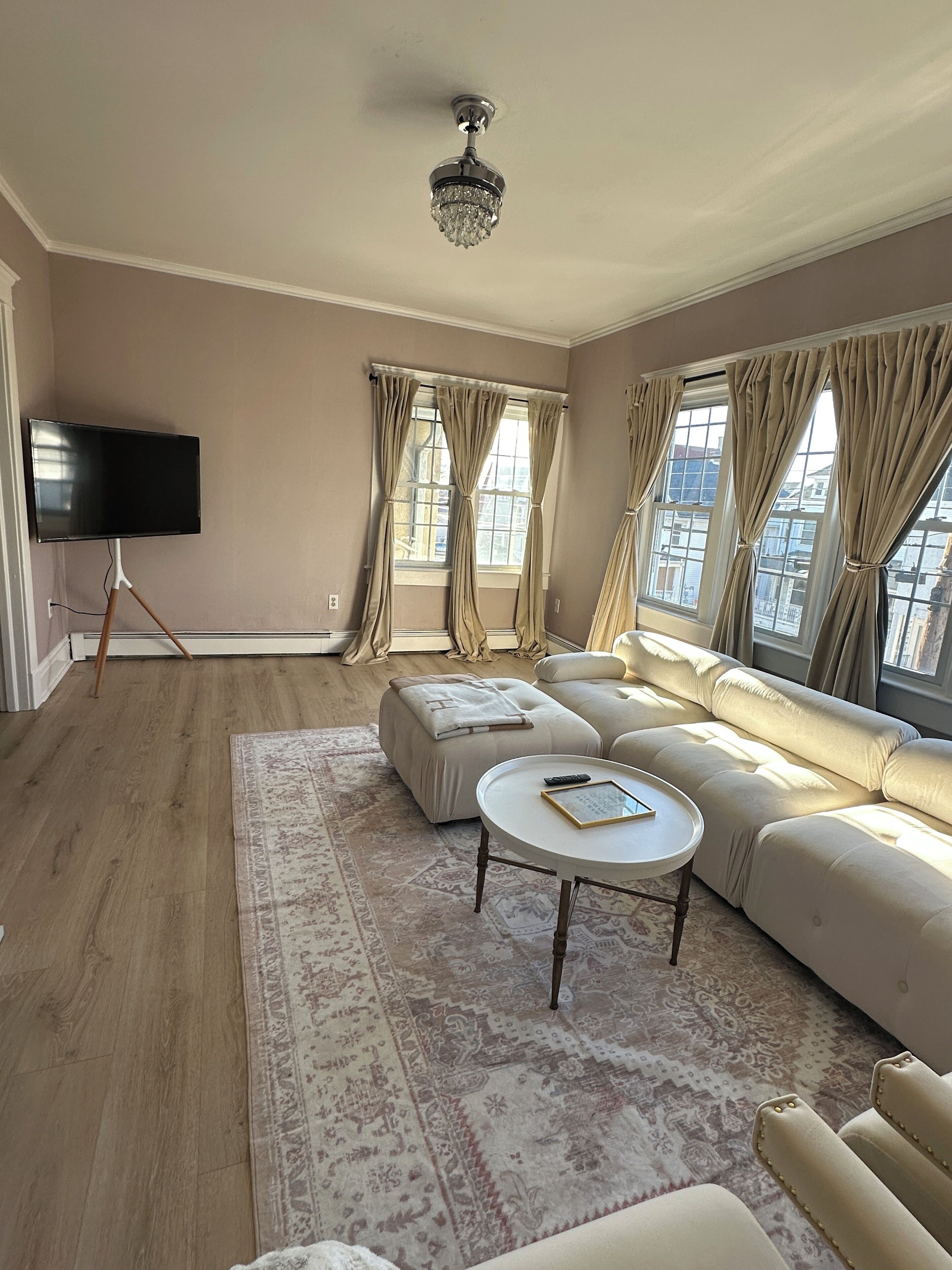 A living room with a large beige sofa, a small round table, a flat-screen TV on a stand, and a patterned rug.